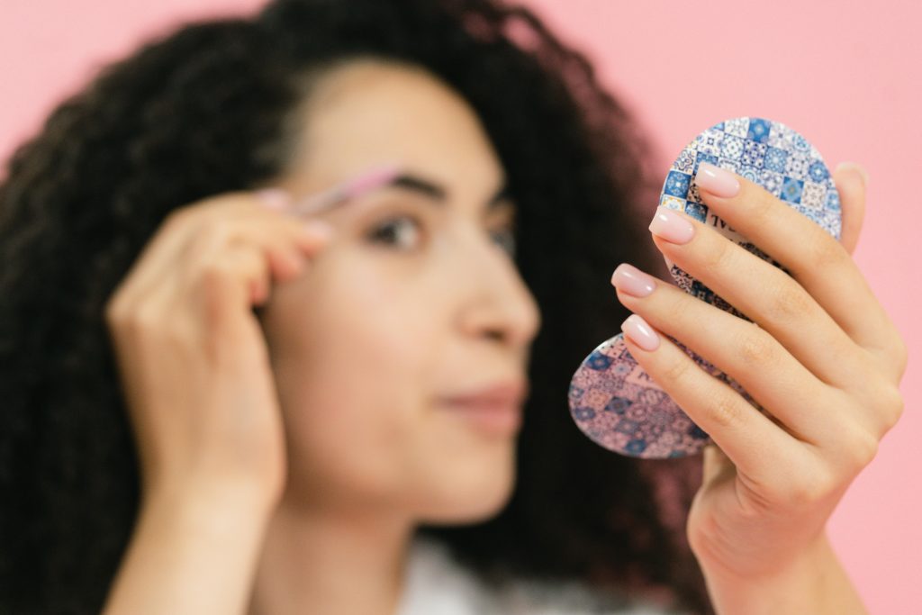 woman using a razor to shave her eyebrow while looking in a mirror